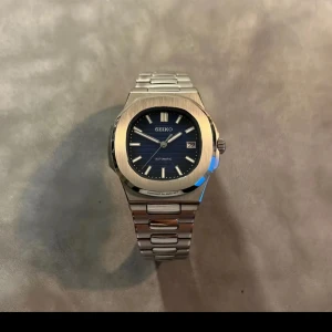 Seiko automatisk klocka med blå urtavla - Snygg Seiko herrklocka i silverfärgat stål med blå urtavla och datumvisning. Klockan har automatiskt urverk som syns genom baksidan och länkarmband i borstat stål. Perfekt accessoar för dig som gillar klassisk design.