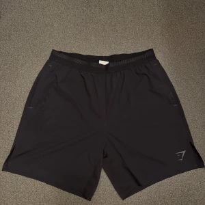 Svarta träningsshorts från Gymshark - Svarta träningsshorts från Gymshark med elastisk midja och diskret logga på benet. Lätta och bekväma med normal passform, perfekta för gymmet eller löpning. Tillverkade i syntetmaterial för bra andningsförmåga.