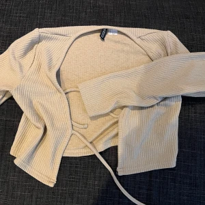 Beige tunnkofta - Säljer en beige ribbad tunnkofta. Modellen är croppad med långa ärmar och knytband 