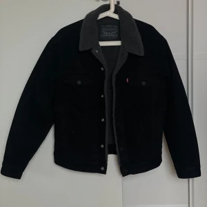 Svart jeansjacka med teddyfoder från Levi's - Säljer en svart jeansjacka från Levi's med mysigt teddyfoder och krage. Jackan har knappar framtill, två bröstfickor och klassisk jeansjacke-look. Perfekt för kyligare dagar och riktigt snygg till vardags.