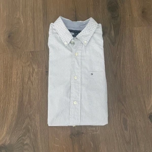 Vit kortärmad skjorta från Tommy Hilfiger - Snygg vit skjorta från Tommy Hilfiger med diskret mörkblått mönster och klassisk button-down krage. Kortärmad modell med små knappar framtill och broderad logga på bröstet. Perfekt till sommarens alla tillfällen.