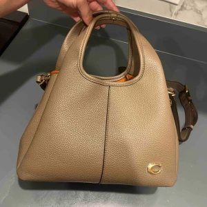 Coach Lori Shoulder Bag - Elegant väska från Coach i en lyxig taupe-nyans med gulddetaljer ✨ Tillverkad i mjukt grained läder och med praktiskt inre fack  perfekt för vardag och jobb! I nyskick, aldrig använd. Justerbar axelrem och medföljande Coach-tag.  💼 Modell: Coach Lori Shoulder Bag 📦 Skick: Som ny 👜 Original tag & rem ingår  📍 Skicka DM vid intresse eller för fler bilder!