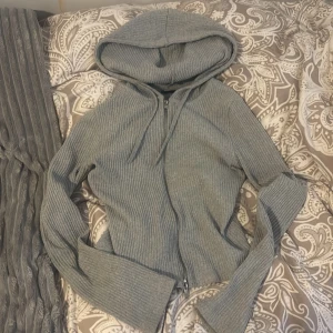 Grå ribbad hoodie från Gina Tricot - Säljer en grå ribbad hoodie från Gina Tricot med dragkedja framtill och huva. Tröjan har långa ärmar och är i ett mjukt, stretchigt material. Perfekt för en avslappnad och stilren look.