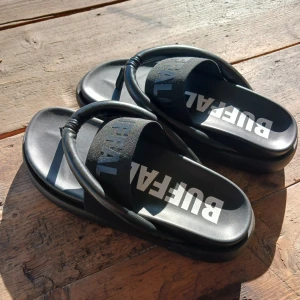 Svarta sandaler från Buffalo - Säljer ett par svarta sandaler från Buffalo med bred rem över foten och tydlig logga i vitt. Skorna är i helt nytt skick och har aldrig använts. Nypris: 1200 kr 