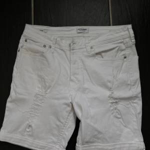 Vita jeansshorts från Jack & Jones - Säljer ett par vita jeansshorts från Jack & Jones med slitna detaljer på framsidan. Perfekta för sommaren och lätta att matcha med olika stilar. 