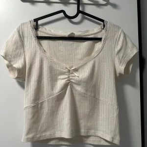 Vit croppad topp med spetsdetaljer  - Söt vit croppad topp i 100% bomull. Toppen har korta ärmar, spetskant vid halsringningen och en liten rynkdetalj framtill. Perfekt för varma dagar och enkel att matcha med jeans eller kjol.