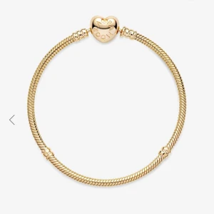 Guldfärgat armband med hjärtformad lås från Pandora - Säljer ett elegant armband från Pandora i guldfärg med ett hjärtformat lås. Armbandet har en klassisk ormkedja och låset är dekorerat med Pandora-loggan. Perfekt att bära som det är eller att dekorera med berlocker.