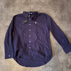 Mörkblå skjorta från Polo Ralph Lauren linne - Snygg mörkblå skjorta från Polo Ralph Lauren med klassisk krage och vita knappar. Skjortan har lång ärm och den ikoniska broderade loggan på bröstet. Perfekt för en stilren look. Passar även L, Linné skjorta