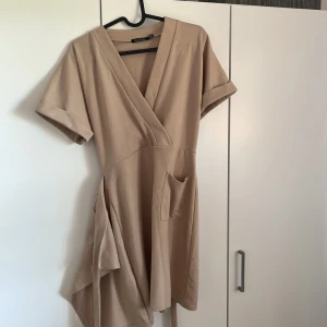 Beige omlottklänning med knytband - Snygg beige omlottklänning med kort ärm och v-ringning. Klänningen har ett brett knytband i midjan och fickor framtill🤎