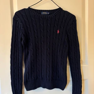 Tröja Ralph Lauren - Säljer en klassisk mörkblå kabelstickad tröja från Polo Ralph Lauren med rosa broderad logga på bröstet i storlek XS. Ganska liten i storleken.