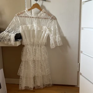 Vit spetsklänning med volanger - Säljer en vit klänning i spets med volangdetaljer och trekvartsärm. Klänningen har rund halsringning och är genombruten i materialet, vilket ger en luftig känsla. Perfekt för studenten eller sommaren!