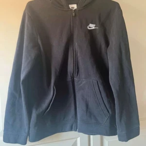 Svart zip-hoodie från Nike - Hej! Jag säljer en svart hoodie med dragkedja från Nike då den är för liten. Klassisk design med vit Nike-logga broderad på bröstet och två fickor framtill. Väldigt skön eftersom den har en mjuk insida.   Obs! XL i barnstorlek, därför har jag satt XS som storlek!!  Vill du veta något mer eller vill ha fler bilder så är det bara att skriva! Priset är såklart inte hugget i sten!