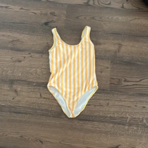 Gul och vit randig baddräkt - En söt baddräkt med gula och vita vertikala ränder. Klassisk modell med breda axelband och rundad halsringning. Perfekt för strand eller poolhäng.