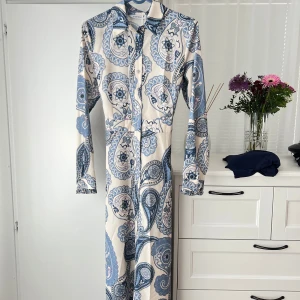 Maxiklänning med bälte - Säljer en långklänning med blått och vitt paisleymönster. Klänningen har lång ärm, krage och knäppning framtill samt ett  bälte i midjan. Materialet känns mjukt och lätt, perfekt för sommaren, hör av er vid intresse💞