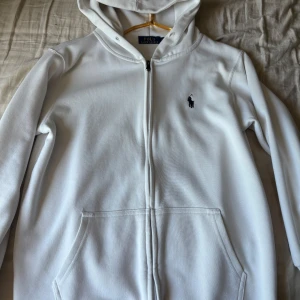 Vit hoodie från Polo Ralph Lauren - Säljer en klassisk vit hoodie från Polo Ralph Lauren med dragkedja och huva. Tröjan har två fickor framtill och den ikoniska loggan broderad på bröstet. Perfekt för en stilren och avslappnad look.