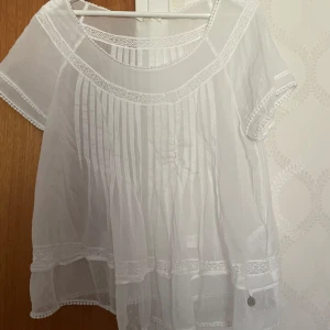 Vit blus med spetsdetaljer från MQ, 365 - Säljer en luftig vit blus från MQ,  365 med fina spetsdetaljer och korta ärmar. Blusen har rund halsringning, dekorativa veck framtill och små prickar i tyget nedtill. Perfekt för vår och sommar! Volang! I nyskick! 