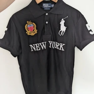 Svart chief keef piké NEW YORK - Svart 100% bomull chief keef piké t-shirt med krage och knappar från Polo Ralph Lauren. Stora broderade detaljer framtill med texten 'NEW YORK'. Stor vit siffra 5 på ryggen och ärmarna. 