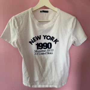 Vit t-shirt med New York-tryck från Zara - Vit t-shirt från Zara med svart tryck där det står 'New York 1990'. Klassisk rund halsringning och korta ärmar. Perfekt för en avslappnad och trendig look. Liten i storlek, skulle säga att den är mer lik S i storlek