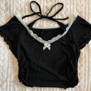 Svart croppad topp med spets och rosett.  - Säljer en svart croppad topp med vit spetskant och liten rosett framtill. Toppen har korta ärmar och knytband i nacken. Jätte söt och fint skick! Jag säljer den eftersom den är för liten för mig. Fråga om ni undrar något!