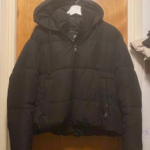 Svart pufferjacka från Zara - Säljer en svart pufferjacka från Zara med huva och dragkedja framtill. Jackan har en oversized passform och är perfekt för kyligare dagar. Snygg och enkel design med dragsko vid huvan.