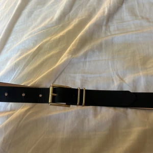 Svart bälte  - Snyggt svart bälte med klassiskt guldspänne och dubbla hållare. Perfekt accessoar för att lyfta din outfit. Bältet har en stilren design och passar till många olika looks.
