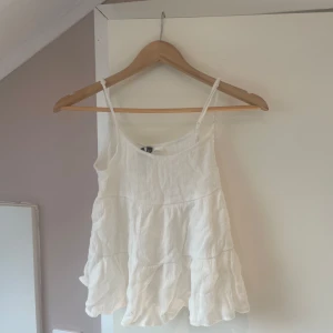 Vit volangig linnetopp - Säljer en söt vit linnetopp med tunna axelband och volangiga lager. Toppen är luftig och perfekt för varma dagar. Passar till både jeans och kjol. Säljer då den är för liten för mig. 