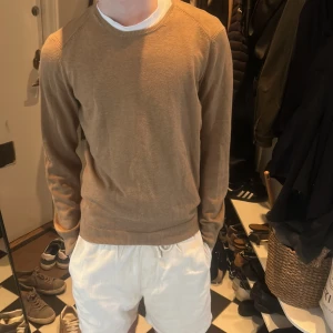 Beige stickad tröja  - Säljer en beige stickad tröja med rund halsringning och bruna mockadetaljer på armbågarna. Tröjan har ribbade muddar vid ärmslut och nederkant. Perfekt för en stilren och avslappnad look. Massimo dutti