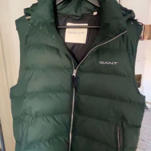 Mörkgrön dunväst från GANT - Snygg mörkgrön dunväst från GANT med dragkedja framtill och två fickor med dragkedja. Västen har hög krage och logga broderad på bröstet. Perfekt för dig som vill ha något skön till sommar 