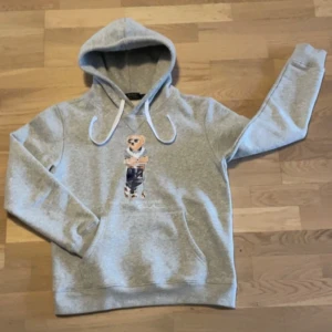 Grå hoodie med Polo Bear från Ralph Lauren - Säljer en grå hoodie från Ralph Lauren med den klassiska Polo Bear-trycket på bröstet. Tröjan har huva med vita snören och en stor magficka. Perfekt för dig som gillar streetwear och vill ha något bekvämt och snyggt.