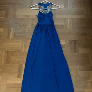 Blå långklänning med dekorerad halsringning - Säljer en blå långklänning med vacker dekorerad halsringning i silver och guldtoner. Klänningen har resår i midjan och är ärmlös med snygg ryggdetalj. Perfekt för högtider och fina tillfällen! Har ett snitt vid sidan! PM för mer bilder!