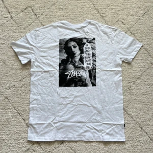 Vit t-shirt med grafiskt tryck från Stüssy - Vit t-shirt från Stüssy med stort svartvitt grafiskt tryck på ryggen och mindre logga på bröstet. Trycket på ryggen föreställer en klassisk konstnärlig bild med textbubbla och Stüssy-logga. Rund hals och korta ärmar. Perfekt för dig som gillar streetwear.