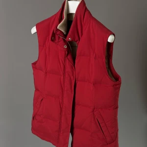 Röd dunväst från Loro Piana - Stilren röd dunväst från Loro Piana med hög krage och diskret broderad logga på baksidan av kragen. Västen har två snedställda fickor framtill och stängs med dragkedja och tryckknappar. Insidan har ett rutigt foder.