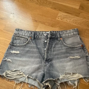Grå jeansshorts med fransar från Zara - Säljer ett par grå jeansshorts från Zara med slitna detaljer och fransar nertill. Shortsen har hög midja, knappar framtill och klassiska fickor både fram och bak. Perfekta för sommaren! De är i storlek 38😊💗