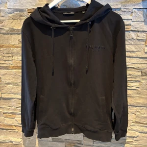 Svart hoodie med dragkedja från Balmain - Säljer en svart hoodie från Balmain Paris med dragkedja framtill och huva. Tröjan har diskret broderad logga på bröstet och stor logga på ryggen. Perfekt för en stilren och avslappnad look.