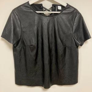 Svart kortärmad topp från H&M - Säljer en svart kortärmad topp i skinnimitation från H&M Divided. Toppen har snygg snörningsdetalj i ryggen och rund halsringning. Materialet är 100% polyester med polyuretanbeläggning för en cool läderlook. Använt två gånger, men det finns en liten defekt.