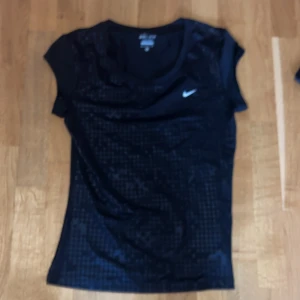 Svart Nike Dri-Fit t-shirt - Svart t-shirt från Nike med diskret mönster och vit logga på bröstet. Tillverkad i Dri-Fit-material som andas, perfekt för träning. Storlek M. 