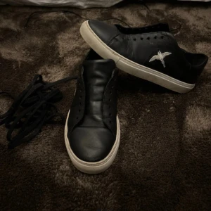 Svarta sneakers med vit sula och broderad fågel - Säljer ett par svarta sneakers med vit sula och en broderad vit fågel på sidan. Skorna har rund tå och platt sula, och är tillverkade i syntetmaterial. Perfekta för dig som gillar stilrena och unika detaljer.   Snören har tagits ut oja rening sätta på efter