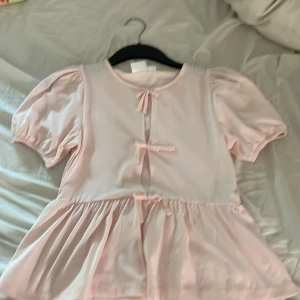 Ljusrosa puffärmad topp från Gina Tricot Young - Supersöt ljusrosa topp med puffärmar och två rosettdetaljer framtill. Modellen har en lös passform och volang nedtill, perfekt för en lekfull och trendig look. Tillverkad i mjuk bomull.
