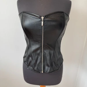 Svart korsett i skinnimitation med dragkedja - Säljer en svart korsett i skinnimitation med dragkedja framtill och snörning baktill. Storlek M och endast använd en gång💕