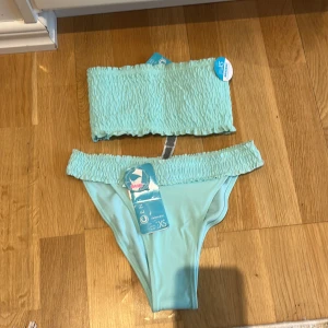 Ljusblå bandeau bikini från CEN - Trendig ljusblå bikini med bandeau-topp. Båda delarna har  detaljer upptill. Man får även med armar till överdelen som är bortagsbara. Bekväm passform. Perfekt för strand eller poolhäng. Materialet känns mjukt och stretchigt. Helt ny och helt oanvänd. Storlek XS