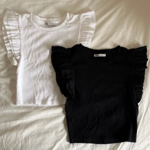 Ribbad topp med volangärmar från Zara - Två ribbade toppar från Zara, en i vitt och en i svart. Båda har korta ärmar med breda volangdetaljer längs axlarna och rund halsringning. Perfekt för att addera lite extra till din outfit. 