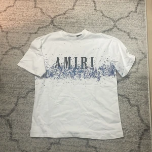 Vit t-shirt från Amiri med blått stänkmönster - Säljer en vit t-shirt från Amiri med stort svart tryck framtill och blått stänkmönster över bröstet och ryggen. T-shirten har rund hals och korta ärmar. Perfekt för dig som gillar streetwear och vill sticka ut med en unik design.
