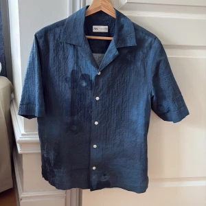 Blå kortärmad skjorta från Zara - Säljer en blå skjorta i linne från Zara med korta ärmar och klassisk krage. Skjortan har knappar framtill och en avslappnad passform, perfekt för varma dagar. Enkel och stilren design som passar till mycket.