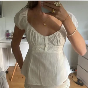 Vit blus med puffärmar och knappar - Säljer en söt vit blus med puffiga korta ärmar och djup urringning. Blusen har knappar framtill och lätt rynkade detaljer vid bysten. Perfekt för dig som gillar romantisk och feminin stil.