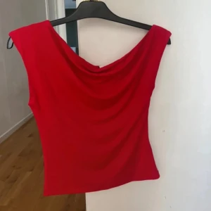 Röd oneshoulder topp - Säljer en snygg röd oneshoulder topp med draperad halsringning. Perfekt för dig som vill sticka ut med en färgstark och trendig look. Toppen är ärmlös och har en smickrande passform.