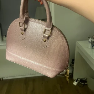 Glittrig rosa handväska - Säljer en superfin glittrig rosa handväska med rundad form och två handtag. Väskan har guldfärgade metalldetaljer och dragkedja upptill. Perfekt för att lyfta vilken outfit som helst!