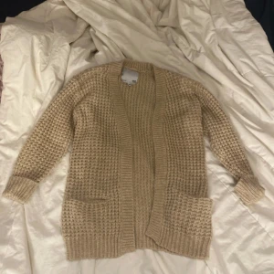 Beige stickad kofta från 157 - Mysig beige stickad kofta från 157 med grovstickat mönster. Koftan har långa ärmar, två stora fickor framtill och breda ribbade muddar vid ärmslut och nederkant. Perfekt för lager-på-lager och enkel att matcha med olika outfits.