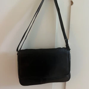 Svart axelväska i skinn - Säljer en stilren svart axelväska i skinn med justerbar axelrem. Väskan har ett klassiskt och minimalistiskt utseende, perfekt för att bära det viktigaste. Passar till många olika outfits och tillfällen.