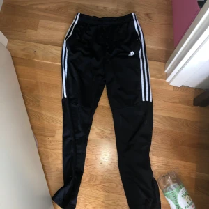 Svarta träningsbyxor från Adidas med vita ränder - Säljer ett par svarta träningsbyxor från Adidas med klassiska vita ränder längs sidorna. Byxorna har dragkedja vid bensluten och elastisk midja. Perfekta för sport eller träning.