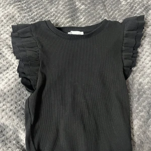 Svart ribbad topp med volangärmar från Zara - Säljer en svart ribbad topp från Zara med snygga volangdetaljer på axlarna. Toppen är ärmlös och har en rund halsringning. Perfekt att styla till jeans eller kjol för en trendig look.